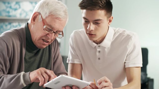Medium shot of senior man in glasses learning how to use tablet computer application, young caregiver helping him