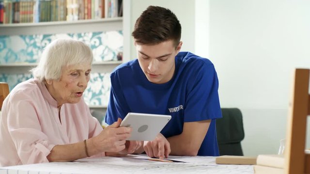 Tracking shot of interested elderly woman sitting at kitchen table and holding tablet computer, young male volunteer sitting next to her and helping her use it