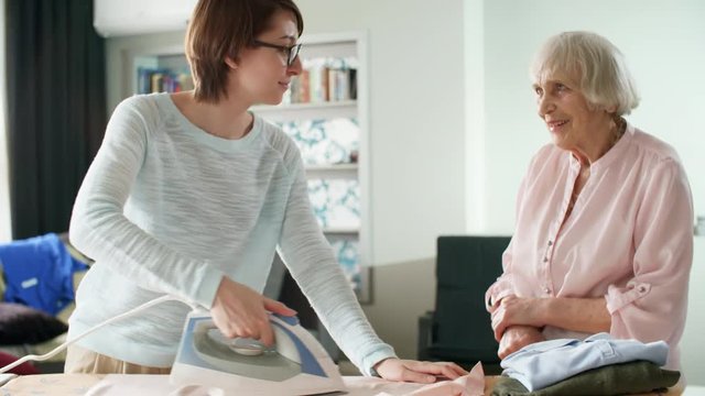 Medium shot of smiling female volunteer ironing clothes on ironing board and talking to elderly woman