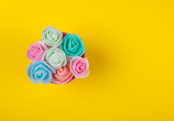 Bunch of multicolored handmade rose buds in a pink bucket on a bright yellow background (minimal...