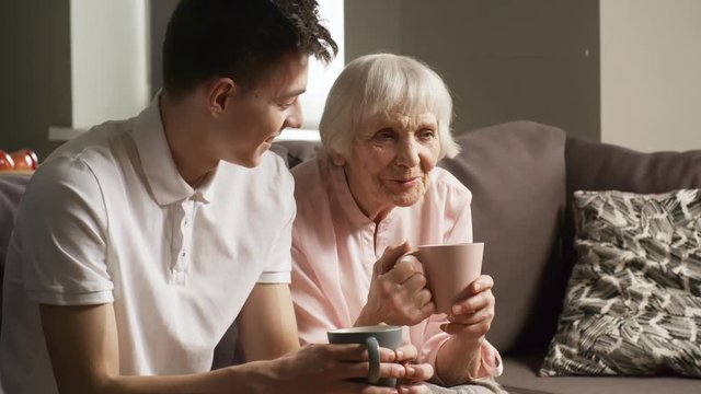 Medium shot of friendly young man smiling and talking to grandmother while sitting on sofa in living room and enjoying tea