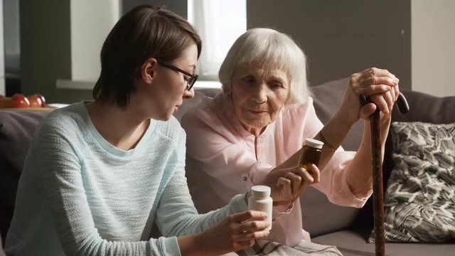 Medium shot of elderly woman sitting on sofa in living room and listening to granddaughter or caregiver, who is explaining doctor&rsquo;s prescription to her