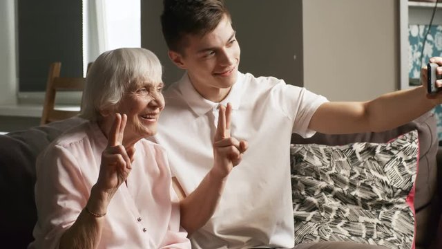 Medium shot of senior woman and her grandson sitting on sofa and making v sign when posing for smartphone camera together