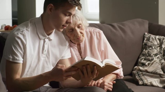 Medium shot of young man sitting on sofa in living room and reading book aloud to grandmother leaning on his shoulder