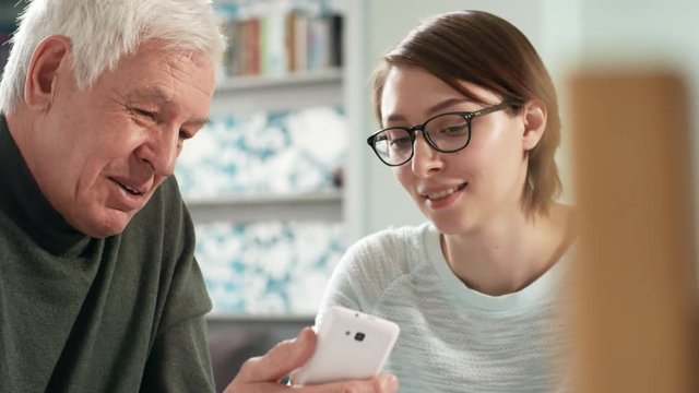 Medium shot of young woman in glasses talking to senior man when teaching him to use smartphone
