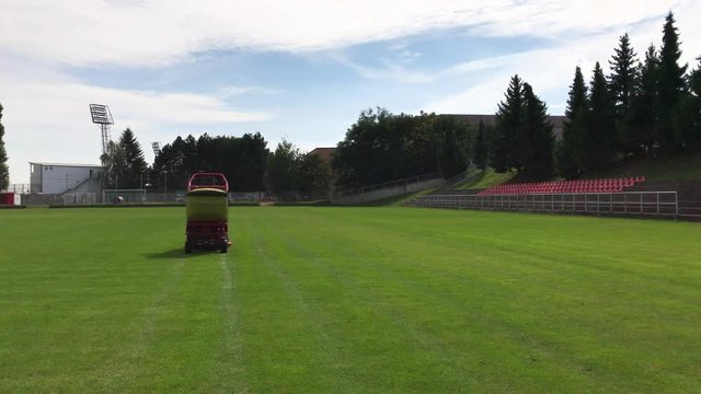 Red and yellow grass-cutting vehicle cuts green grass on football field moving away, lines after grass-mower on green before match