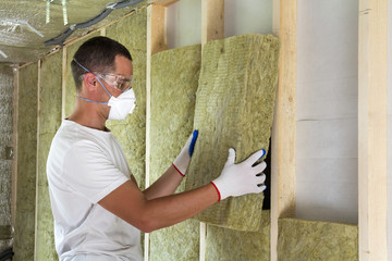 Worker in protective goggles and respirator insulating rock wool insulation in wooden frame for...