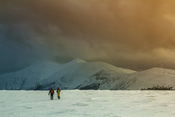Fototapeta na wymiar Winter panorama. Back view of two tourists hikers with backpacks going in snowy valley to magnificent mountain range under gloomy cloudy sky. Active lifestyle, tourism and extreme sports concept.