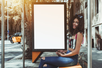 A cheerful curly cute girl is sitting on the metal bench of an urban bus stop, holding smartphone...