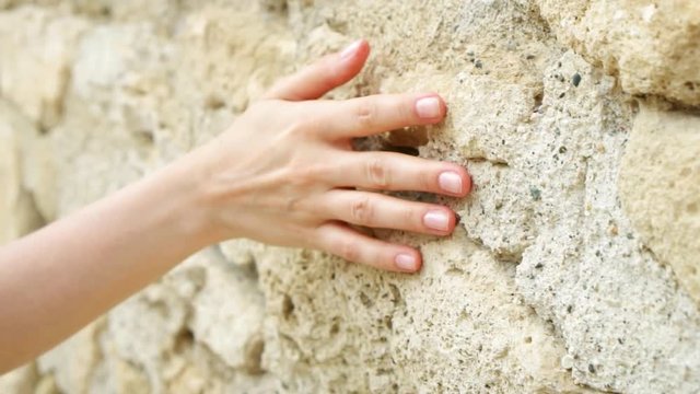 Woman sliding hand against old ancient stone wall in slow motion. Female hand touching hard rough surface of rock on sunny summer day