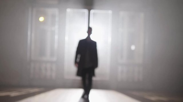 Blurry video imaging showing young short haired man in long dark coat with straight collar walking away from large glowing windows towards camera in empty darken hall.