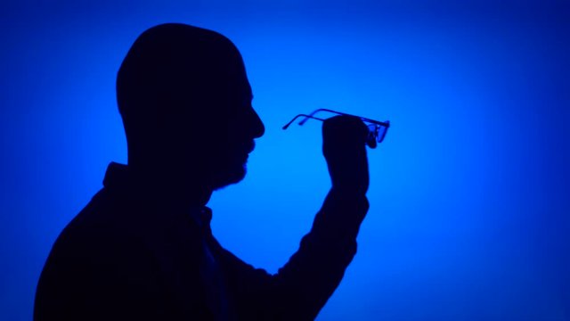 Silhouette of senior man with eyeglasses. Male's face in profile putting on glasses on blue background. Black contour shadow of grandfather's half-face holding spectacles