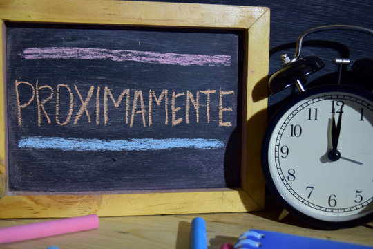 Proximamente in spanish on phrase colorful handwritten on blackboard. Education and business concept. Alarm clock, chalk, books on black background
