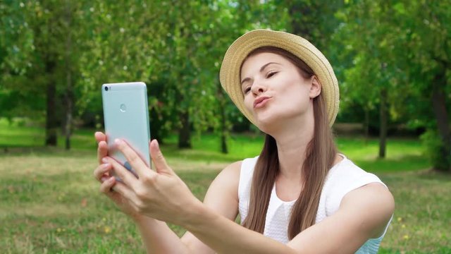 Young female student in white shirt sitting on grass on college campus doing selfie on mobile phone. Tourist in hat make photos on smart phone in public park enjoying summer sunny day