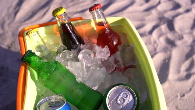 Colorful soda drinks and filled ice cubes in a coolbox on the beach sand. 4K video