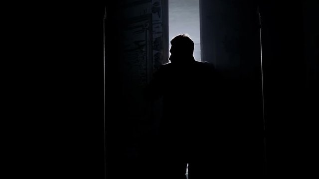 Silhouette of mature man in long dark coat with straight collar opening two massive antique wooden doors revealing empty room in daylight with white dusty walls and big arched windows.