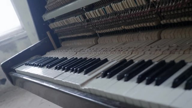 Retro, wooden, brown piano with broken body frame and dusty keys revealing inside hammers and strings is completely abandoned in empty hall surrounded by dirty white walls and large glazed windows