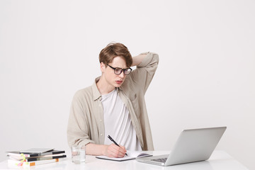 Portrait of thoughtful young man student wears beige shirt and spectacles looks concentrated and study at the table using laptop computer and notebooks isolated over white background