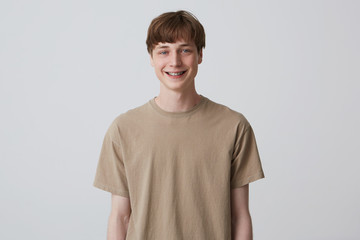 Studio portrait of smiling handsome young man with healthy teeth and metal braces wears beige tshirt and looks directly in camera isolated over white background Feels happy and thinks about girlfriend - obrazy, fototapety, plakaty