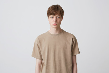 Portrait of serious handsome young man student with short haircut and blond hair in beige t shirt standing and looking directly in camera isolated over white background Feels concentrated