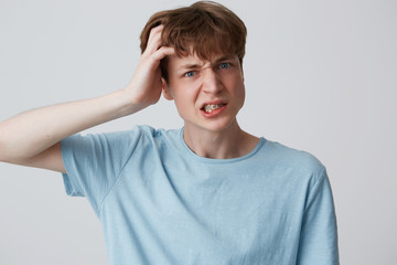 Closeup of angry mad young man with braces on teeth wears blue t shirt keeps hand on head and looks irritated isolated over white background