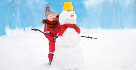 Fototapeta na wymiar Little boy in red winter clothes having fun with snowman in snowy park