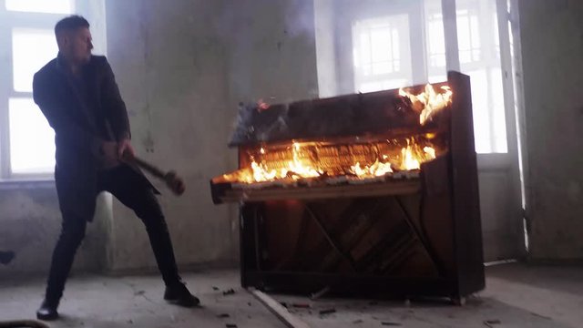 Attractive young bearded man with stylish short haircut dressed in grey coat is agressively hitting flaming classic wooden piano with huge metallic hammer in cold empty room.