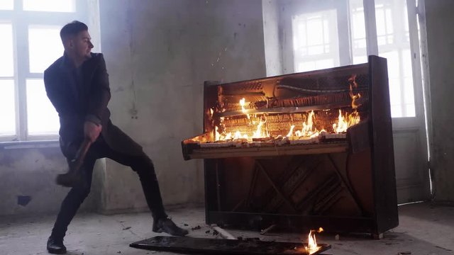 Young bearded man with stylish short haircut dressed in grey coat is agressively hitting flaming obsolete wooden piano with massive metallic hammer in cold empty room.