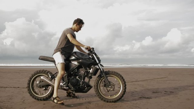 Young handsome hipster man sit on modern custom motorcycle cafe racer on the black sand ocean beach and drive away near the water. Adventure and travel concept. Surfing spot with ocean waves.