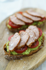 Pork tenderloin sandwich with mashed avocado, tomatoes and maldon salt. Served on a wooden cutting board, high resolution.