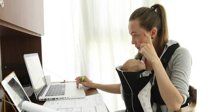 Multitasking mom working, nursing newborn baby. Mother holding child at ergo backpack home, looking on laptop computer. Businesswoman nursing son. People, family, maternity