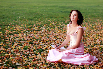 Fine emotional brunette writing on paper, sitting on autumn leaves and grass. Copy space. Yonge girl in vintage pink pastel with live expressions on beautiful country and old background.