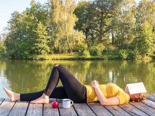 Junge Frau liegt mit Buch auf dem Gesicht auf einenm Holzsteg am See, Ruhe, Stille, Auszeit