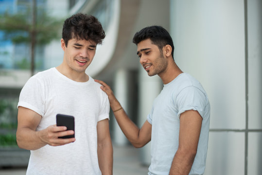 Puzzled men reading online news on smartphone, his Indian friend supporting him and touching his shoulder. Surprised guys viewing message on gadget outdoors. Information concept