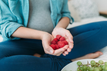 Cropped photo of young pregnant woman eating grapes and raspberries in bed