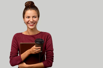 Fototapeta premium A cheerful brunette girl with hair collected in a bundle holds a leather folder and a Cup of coffee in her hand. A positive business lady or student laughs and looks away