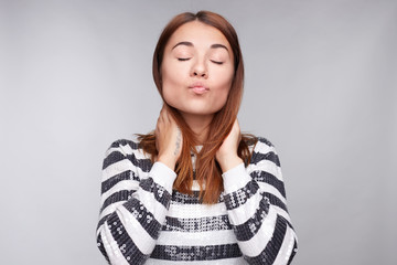 Fashionable young good looking female with dark ginger hair keeps lips round, looks coquette, keeps hands on neck, sends kiss at camera, poses against gray background, having fun and flirting look.