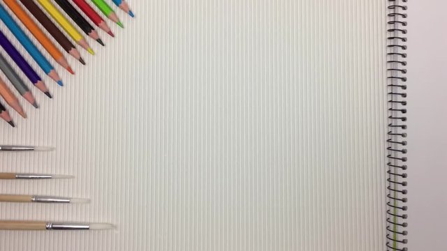 Escena de una mesa con lapices de colores y pinceles, aparece una libreta por la derecha.