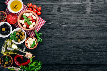 Ingredients for Italian caprese salad. Mozzarella cheese, cherry tomatoes, basil leaves, olives, oil, pepper. On a black wooden background. Free space for text.