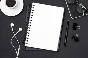 Business flat lay composition: desk with notebook, pencil, coffee cup, glasses, headphones and supplies on black table. Note pad space for text input. Top view shot. Businessman or student desk.