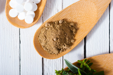 herbal leaves, ground herb powder and medicament pills on bamboo spoons, white wooden table