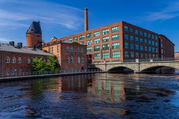 red-brick buildings on the shore of a rapids flowing through the city, backdrop of a blue sky reflecting on the surface of the rapids