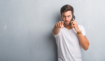 Handsome young man over grey grunge wall speaking on the phone pointing with finger to the camera and to you, hand sign, positive and confident gesture from the front