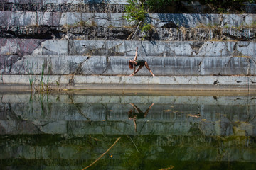 A red-haired young woman in green overalls is engaged in yoga at the degree of an abandoned marble quarry. In the water of a flooded marble quarry the reflection of a girl engaged in pilates