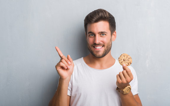 Handsome young man over grey grunge wall eating chocolate chip cooky very happy pointing with hand and finger to the side