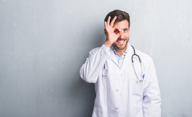 Handsome young doctor man over grey grunge wall doing ok gesture with hand smiling, eye looking through fingers with happy face.