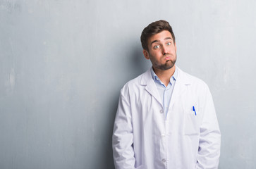 Handsome young professional man over grey grunge wall wearing white coat puffing cheeks with funny face. Mouth inflated with air, crazy expression.