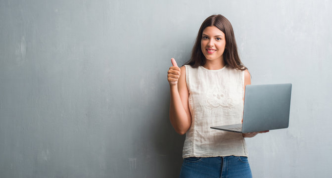 Young brunette woman over grunge grey wall using computer laptop happy with big smile doing ok sign, thumb up with fingers, excellent sign