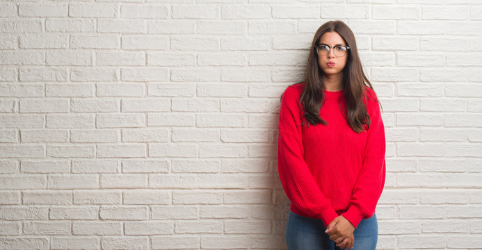 Young brunette woman standing over white brick wall puffing cheeks with funny face. Mouth inflated with air, crazy expression.