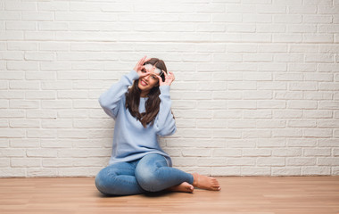 Young brunette woman sitting on the floor over white brick wall with happy face smiling doing ok sign with hand on eye looking through fingers